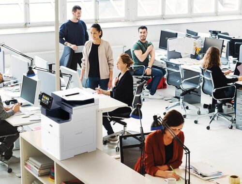 Un groupe de professionnels collaborant dans un bureau, utilisant des ordinateurs et l'imprimante multifonction Xerox® VersaLink® B625 pour leurs tâches.